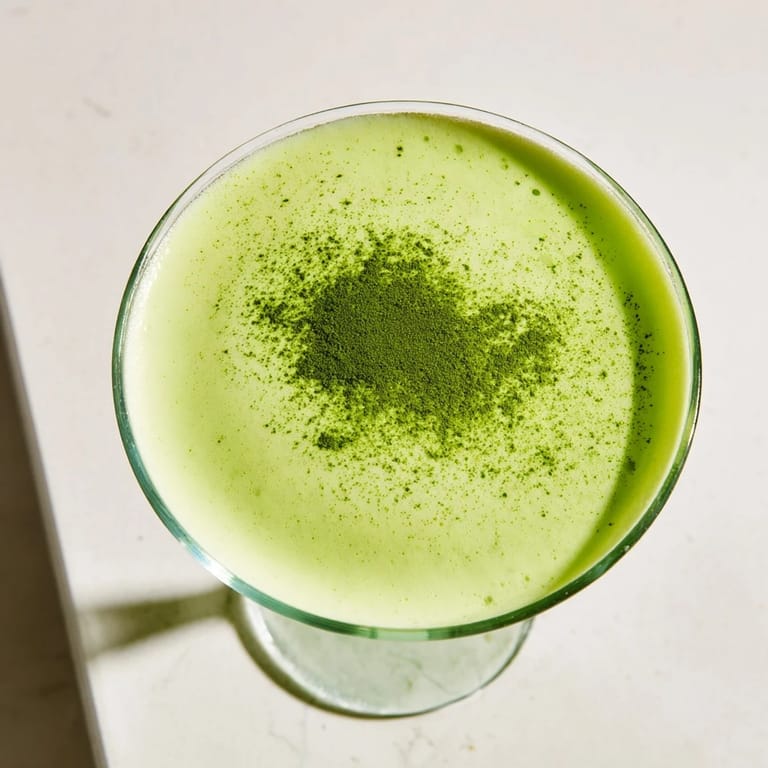 A Matcha Martini cocktail is garnished with white chocolate shavings, beside a cocktail shaker and matcha powder.