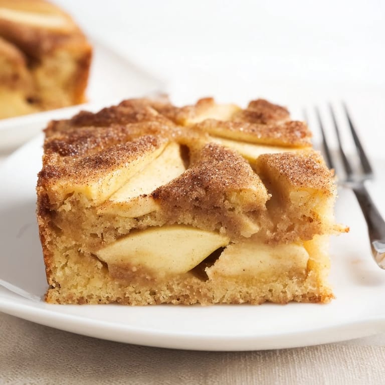 Freshly baked Dutch-style apple cake in a dish, topped with cinnamon sugar and served warm with a dollop of whipped cream.