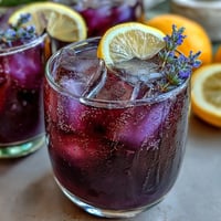 A refreshing purple lemonade with lavender, citrus, and grape juice, served over ice with lemon slices.
